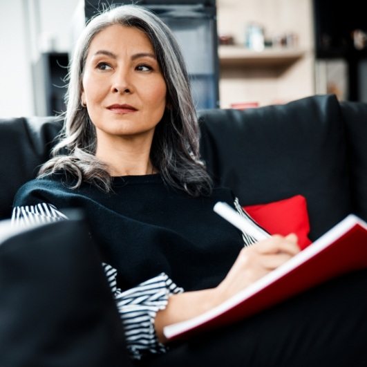 middle aged lady considering dental implant tooth replacement writing in notebook on black couch